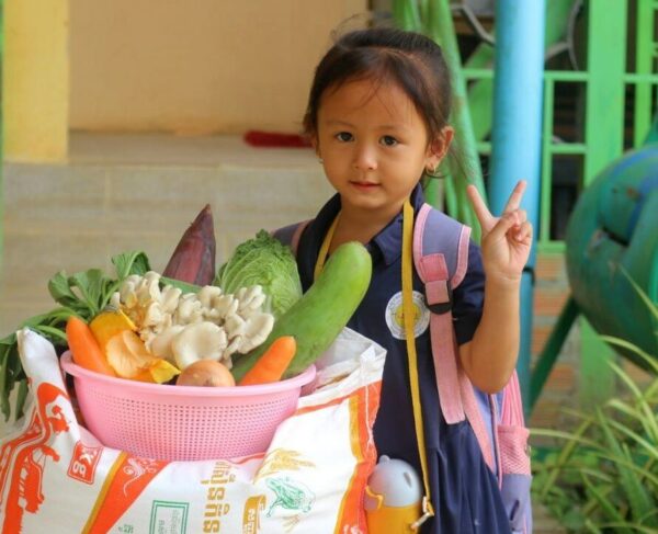 Little girl stands with a basket of vegetables and large bag of rice