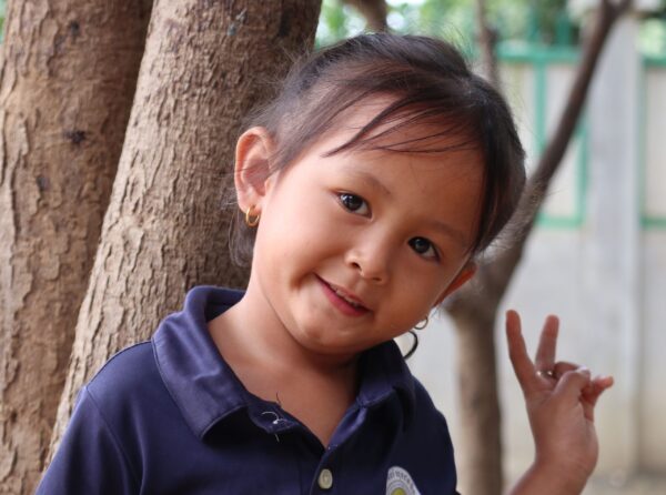 Little girl in blue shirt makes peace sign in front of a tree